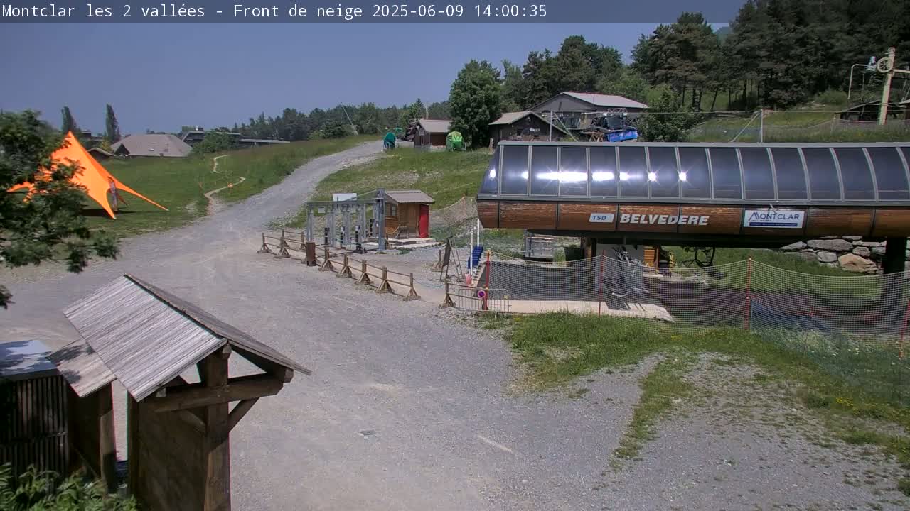 A chairlift station sits on a gravel path amidst green grass and trees under a clear blue sky.