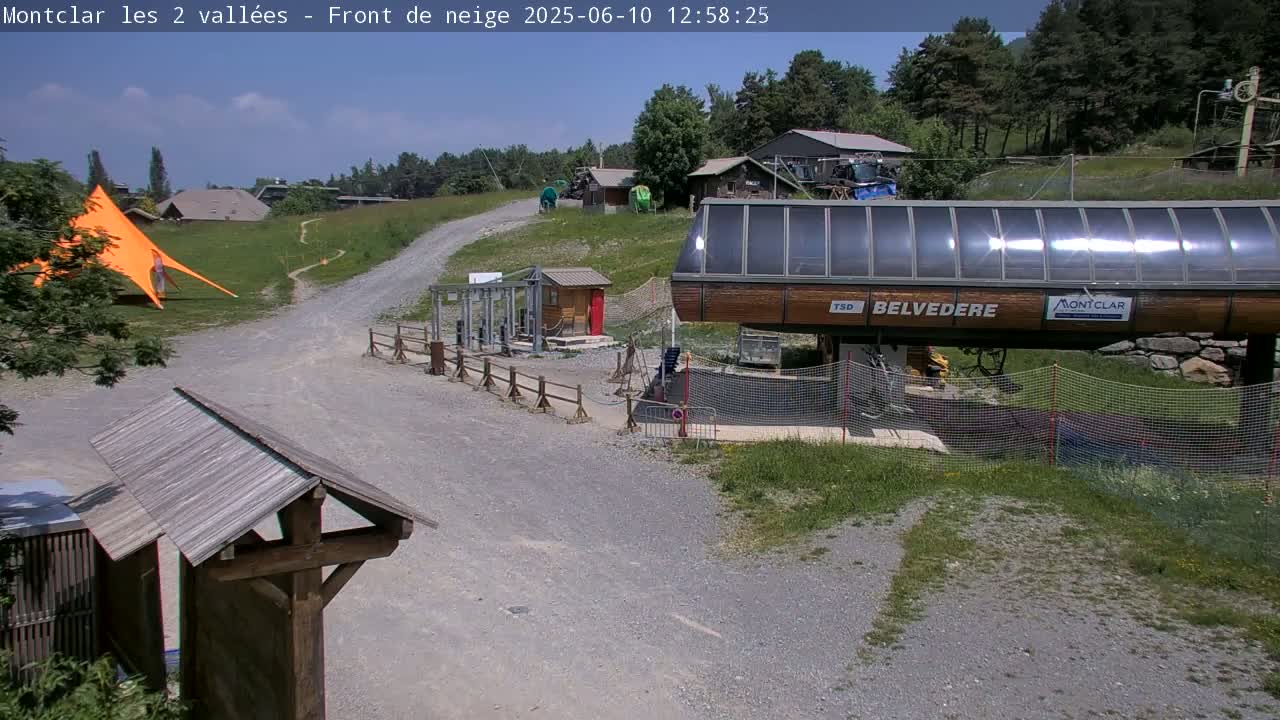 A chairlift base station sits at the end of a gravel path in a sunny, mountainous area with green grass and trees.