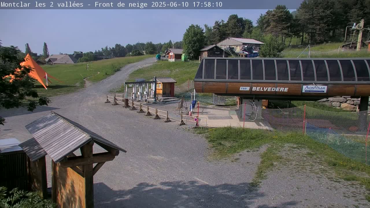 A chairlift station sits on a gravel area surrounded by green grass and trees under a clear, sunny sky.