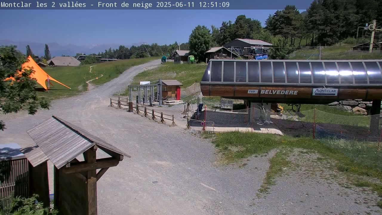 A sunny outdoor scene shows a gravel path leading to a chairlift station, with a large orange canopy and some buildings in the background.