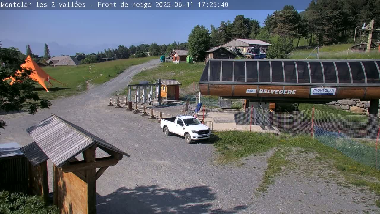 A sunny outdoor scene shows a chairlift station, a white pickup truck, and a large orange tent on a grassy area with gravel paths, surrounded by trees and buildings.