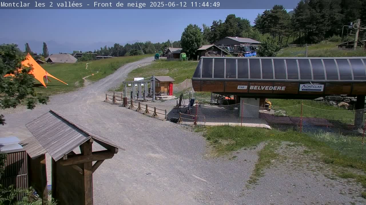 A chairlift station and surrounding area on a sunny day, with a gravel path leading to it and grassy areas nearby.
