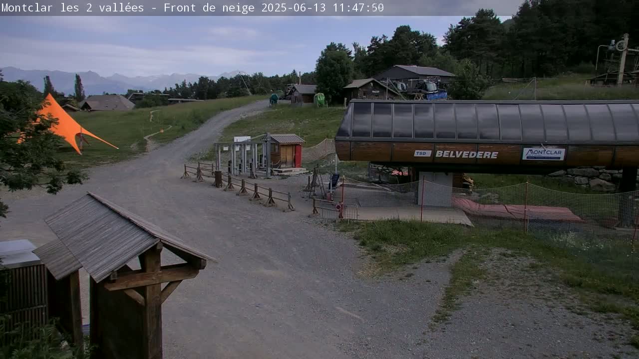 A gravel path leads to a chairlift station in a mountainous area under partly cloudy skies, with an orange canopy and various buildings in the background.