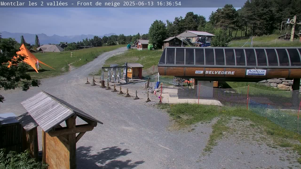 A gravel path leads to a chairlift station in a sunny, mountainous area with green grass and a few buildings.