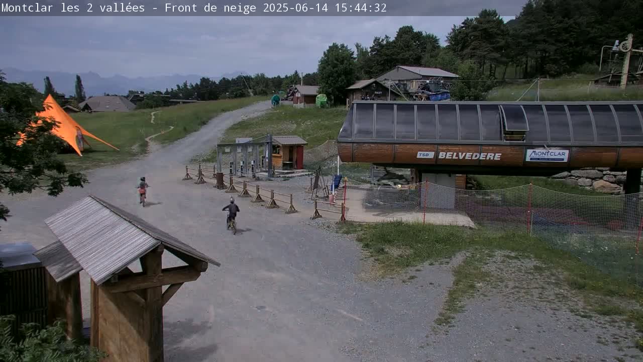 A mountaintop scene under a cloudy sky shows a gravel road with two cyclists, a chairlift, and a large orange canopy.