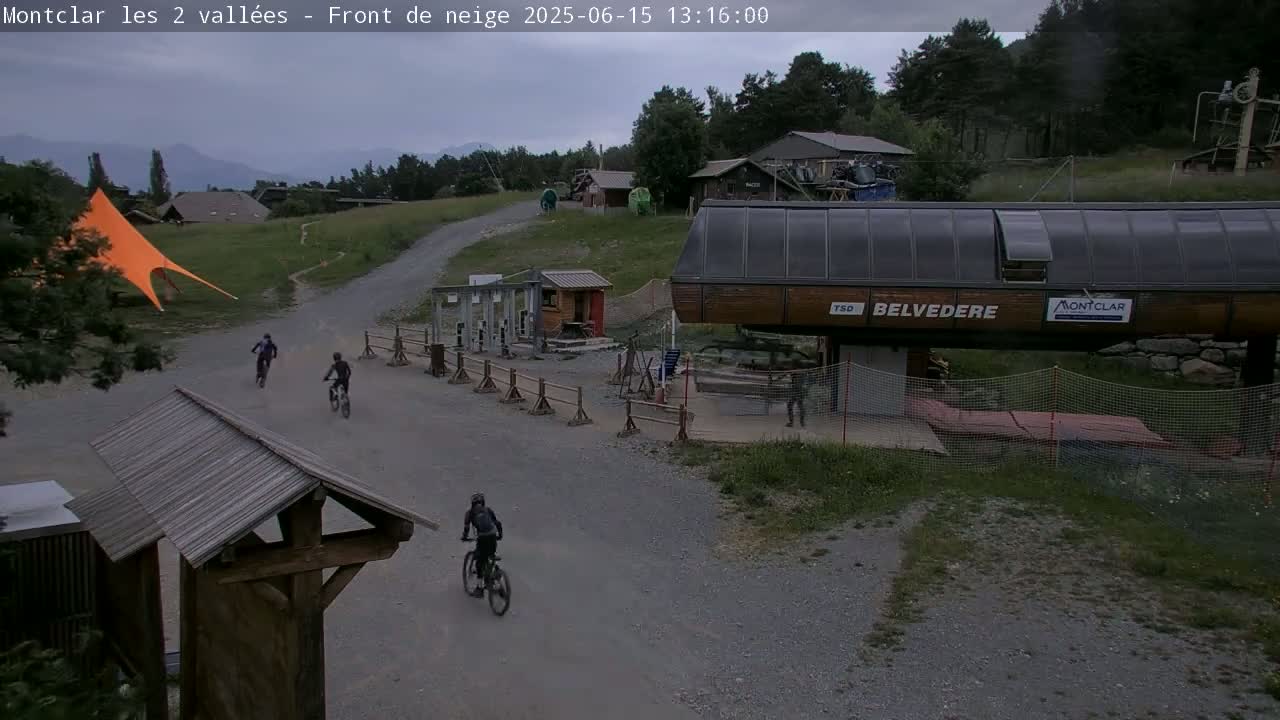 A mountaintop scene under an overcast sky shows three mountain bikers on a gravel path near a chairlift and several buildings.
