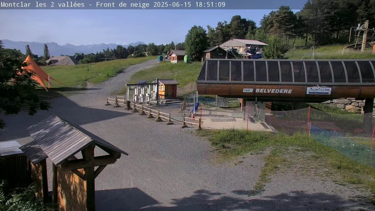A sunny mountaintop scene shows a chairlift station, gravel paths, and sparse vegetation, with mountains in the background.