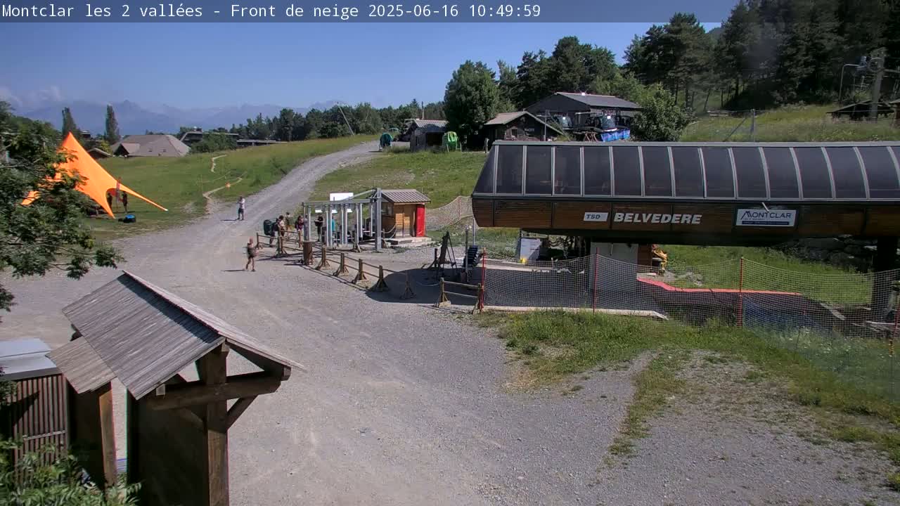 A chairlift station sits on a gravel path in a mountainous area on a sunny day, with several people walking nearby and a large orange tent in the background.