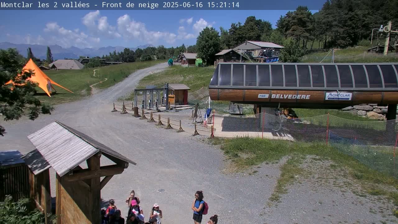 A sunny outdoor scene shows a mountainside ski lift station with several people standing near a gravel path, an orange canopy tent, and a distant mountain range under a partly cloudy sky.
