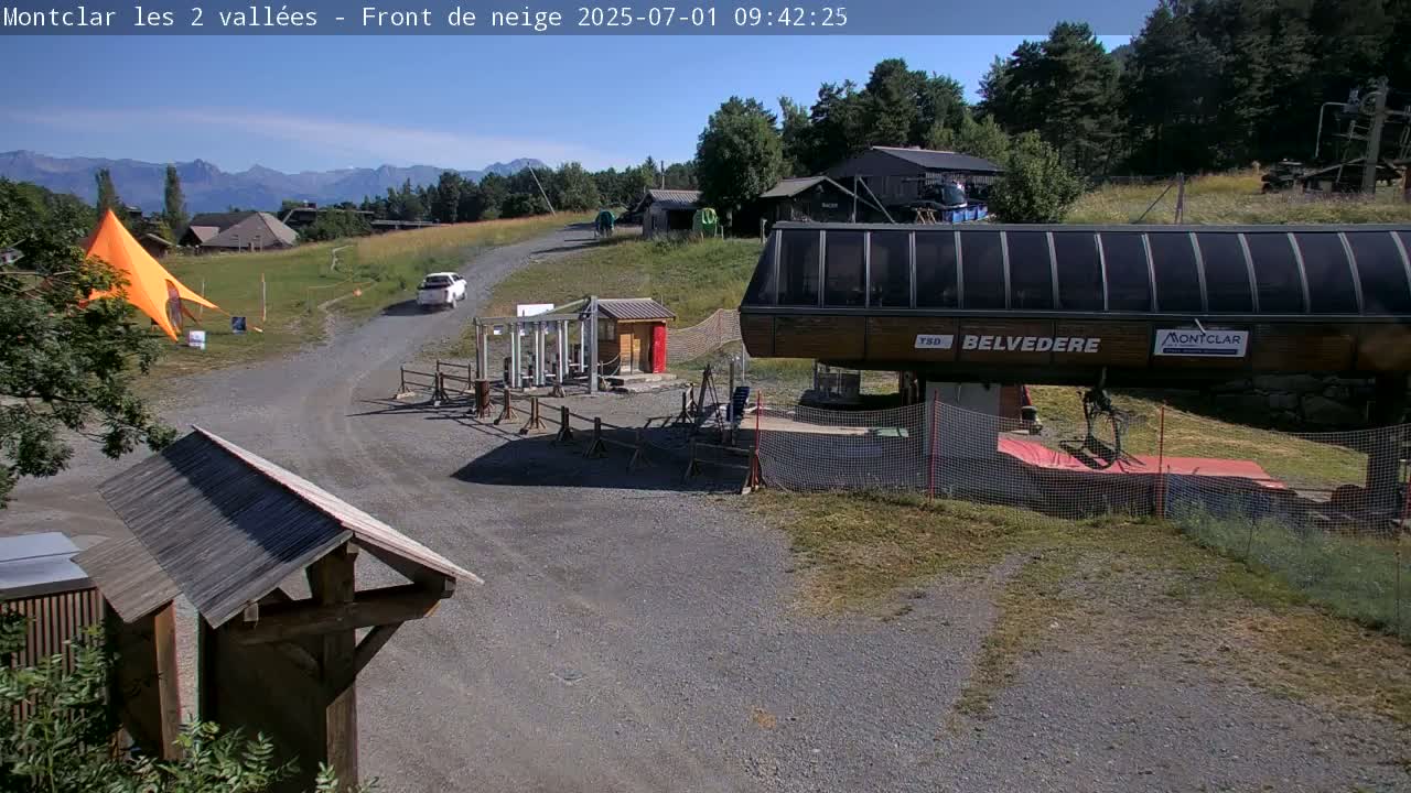 A sunny mountaintop scene shows a gravel road leading to a chairlift, with a car and a large orange tent visible in the background, mountains in the far distance.