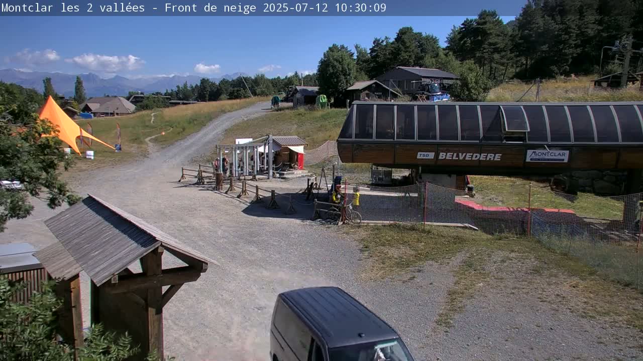 A sunny mountain vista shows a chairlift station, a gravel road, a few buildings, and a bright orange tent, with mountains in the background.
