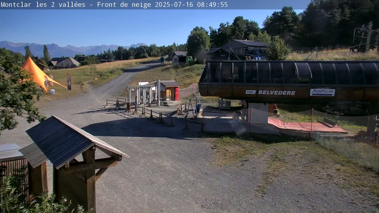 A chairlift base station sits on a gravel path near some buildings and an orange canopy, under a clear sunny sky with mountains in the distance.