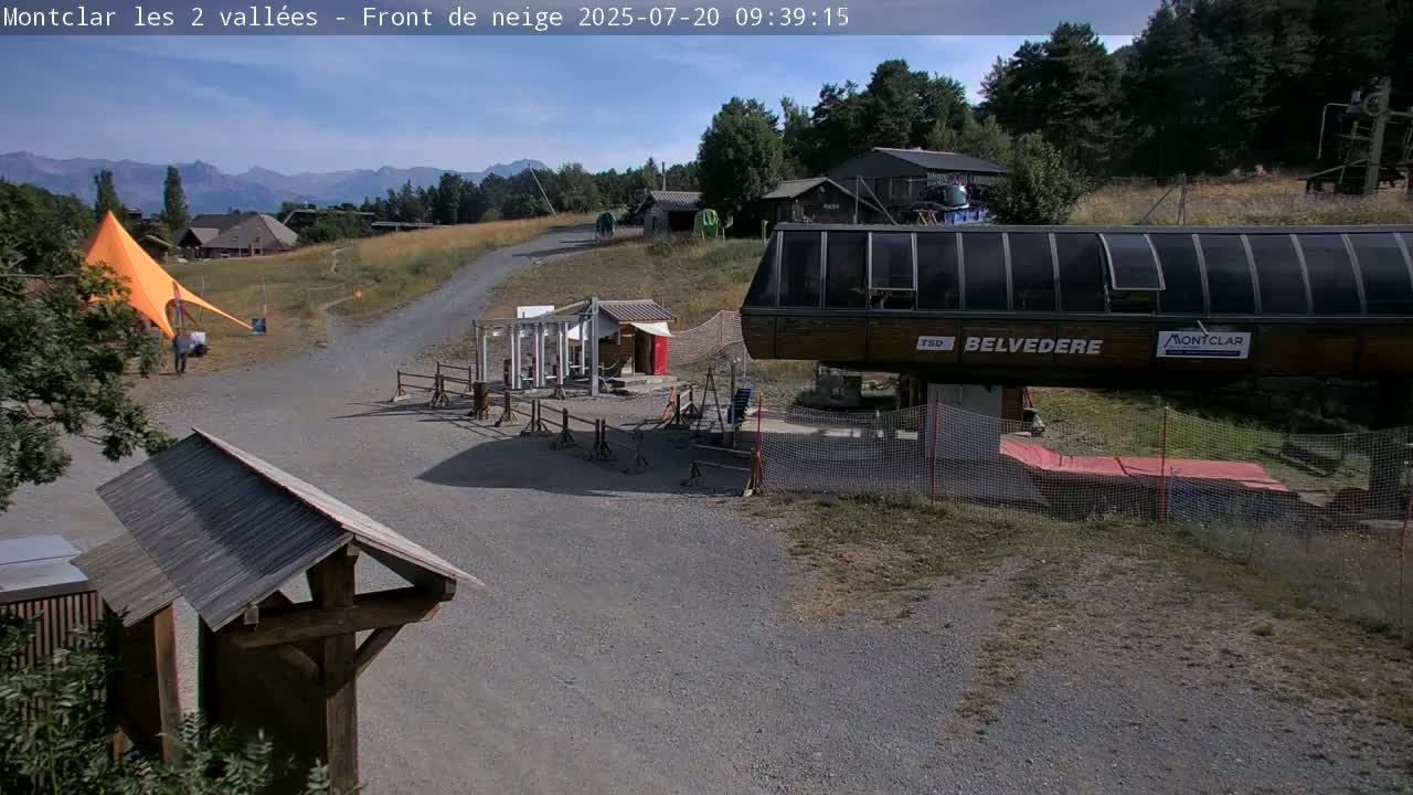 A sunny day reveals a mountain vista, a chairlift station, a gravel path, and a bright orange tent in a grassy area.