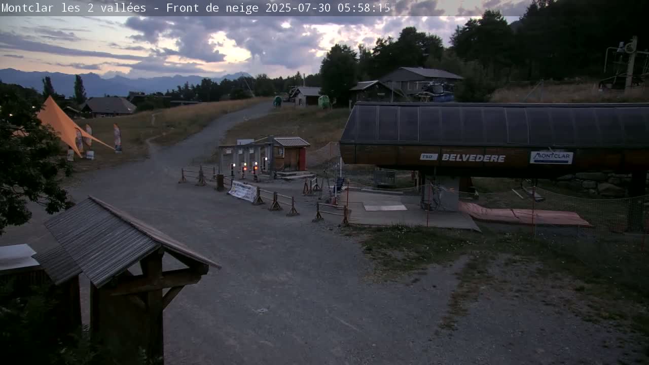 A sunny day reveals a mountain vista, a chairlift station, a gravel path, and a bright orange tent in a grassy area.