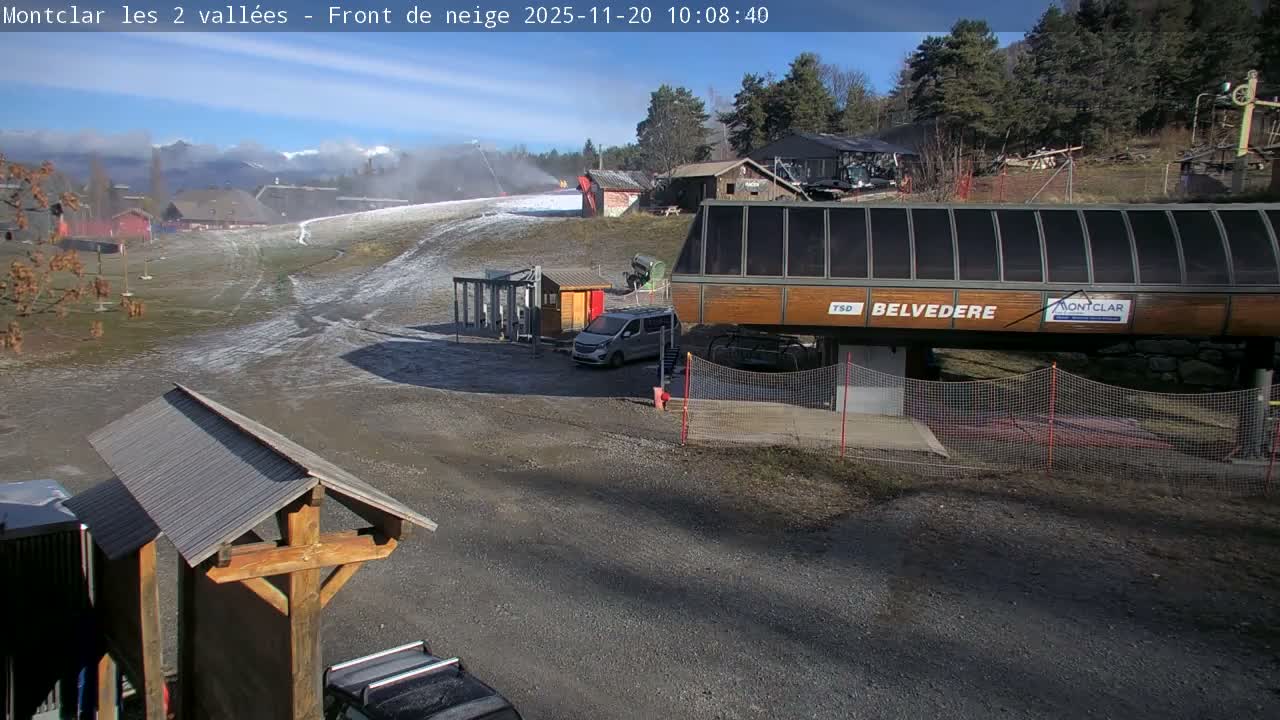Under a bright, sunny blue sky, a ski resort prepares for the season with a snow cannon actively making snow on a partially bare slope, alongside a large ski lift station and a parked van.