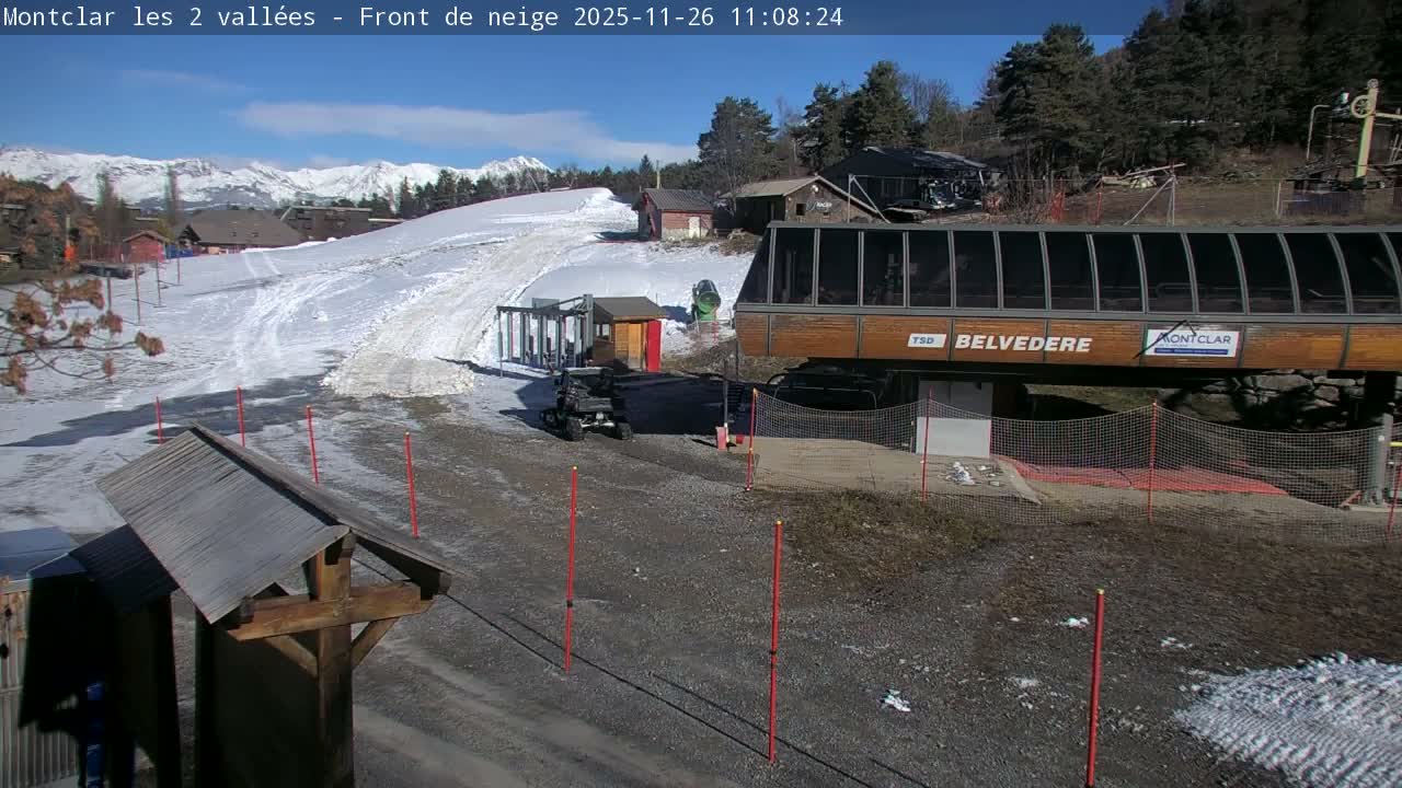 Under a clear blue sky and bright sunshine, a ski resort features a partially snow-covered slope leading to a Belvedere ski lift station and an ATV, with distant snow-capped mountains visible.