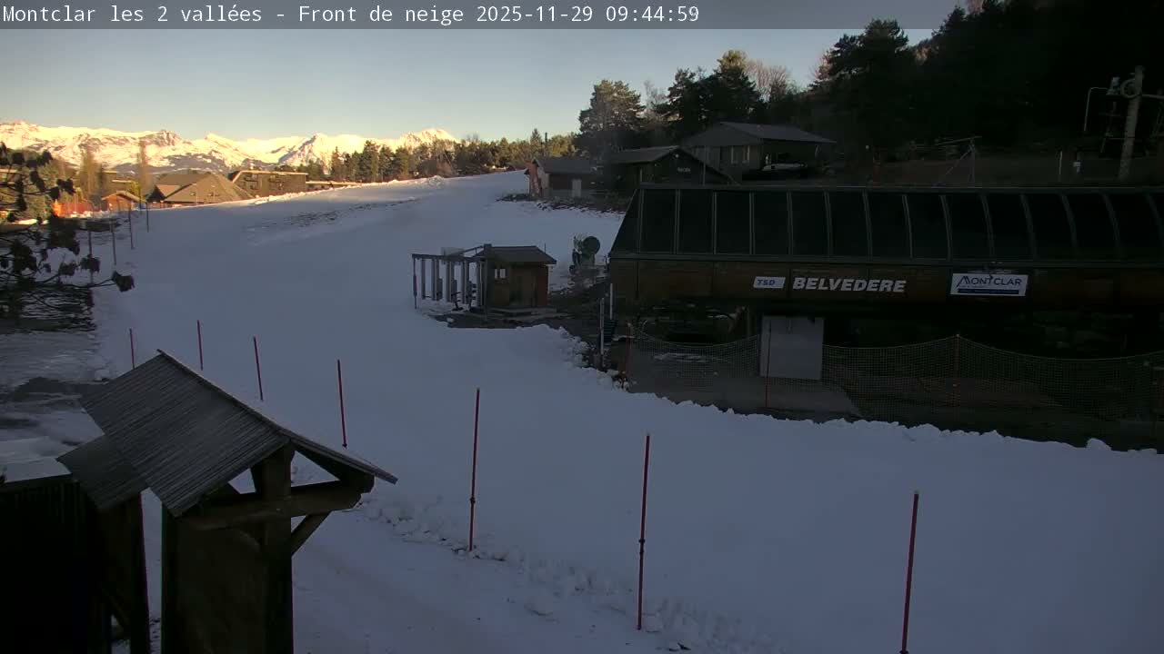 A sun-drenched ski resort features a wide, snow-covered slope leading past buildings and a ski lift station, with distant snow-capped mountains under a clear sky.