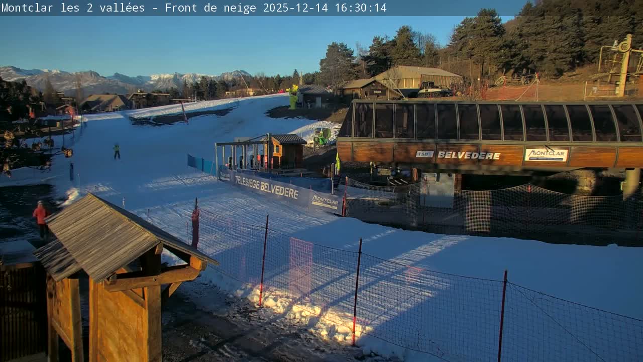 A sun-drenched ski resort features a wide, snow-covered slope leading past buildings and a ski lift station, with distant snow-capped mountains under a clear sky.