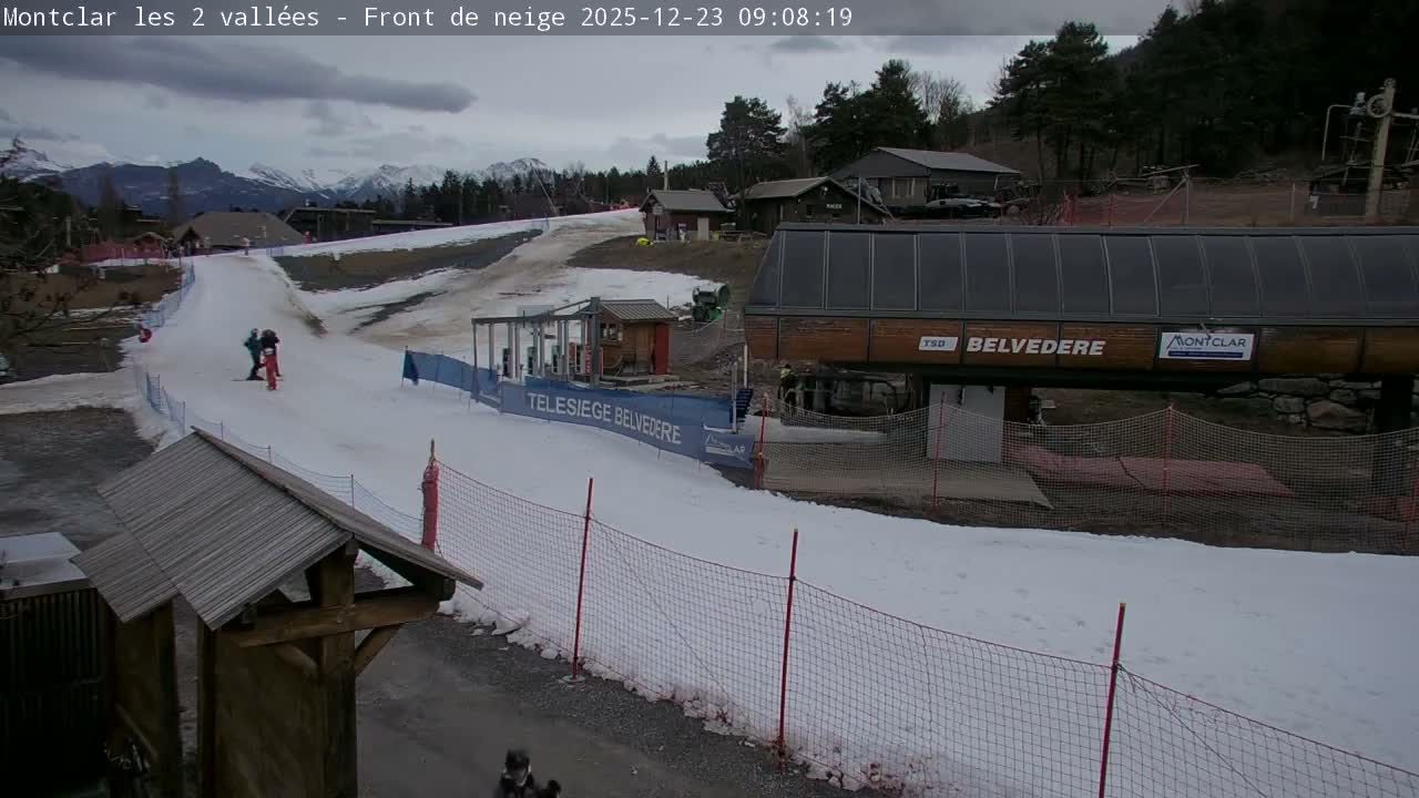 The image displays a snowy ski slope with a few skiers descending, a chairlift station, and mountain ranges in the background under an overcast sky.