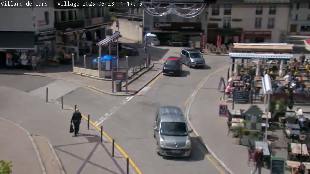 A sunny day in a village square shows several cars, pedestrians, and outdoor seating areas at restaurants.