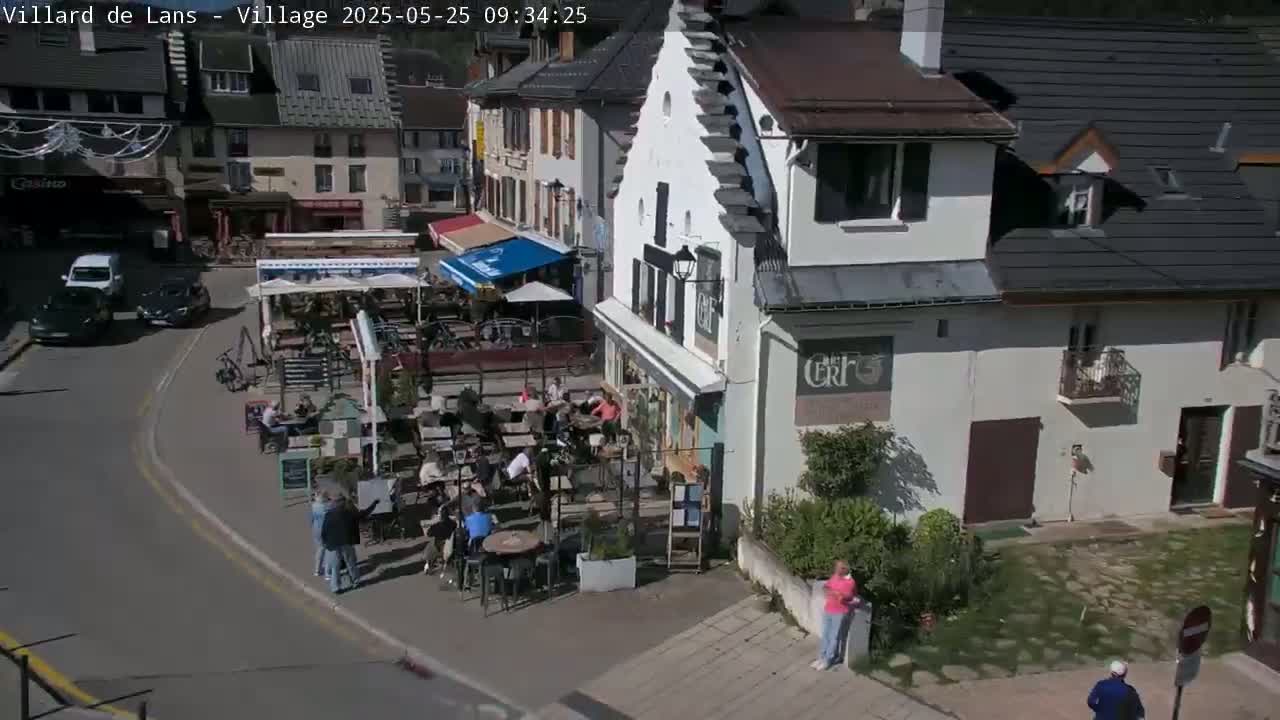 A sunny outdoor scene shows a town square with several cars parked, people sitting at outdoor cafes, and buildings lining the street.