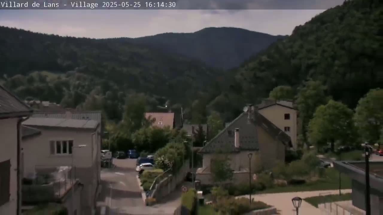 A village nestled in a valley, surrounded by dark green hills under a partly cloudy sky.