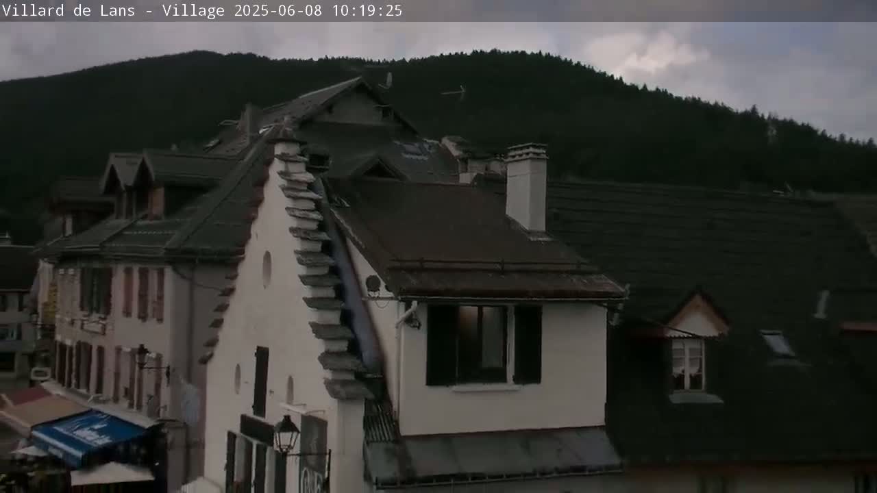 A mountain village under a cloudy sky shows several buildings with dark roofs and light-colored walls.
