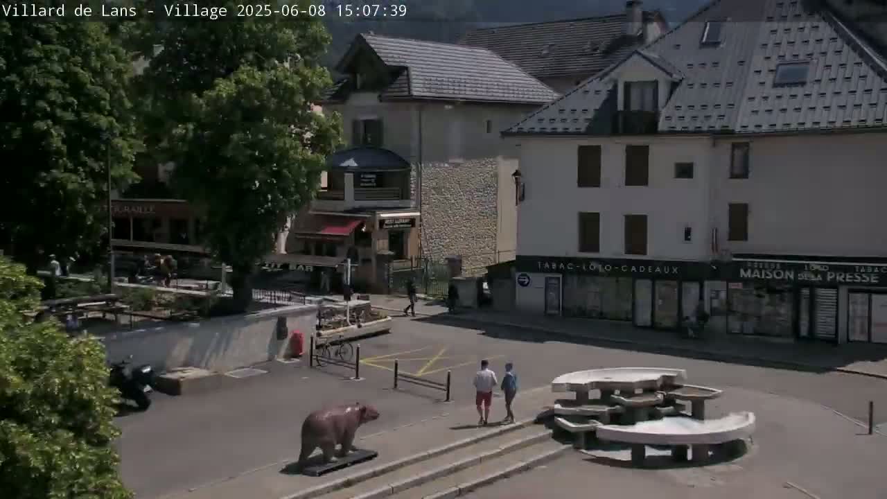 A sunny outdoor scene shows a town square with several buildings, a small fountain, a bear statue, and a few people walking.