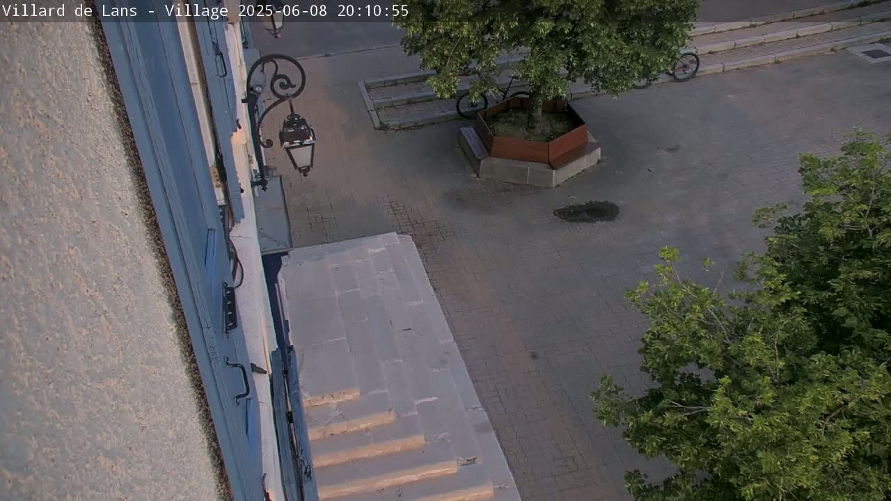 A high-angle view shows a stone staircase leading down to a paved area with a tree in a planter, a bicycle, and some small puddles under a twilight sky.