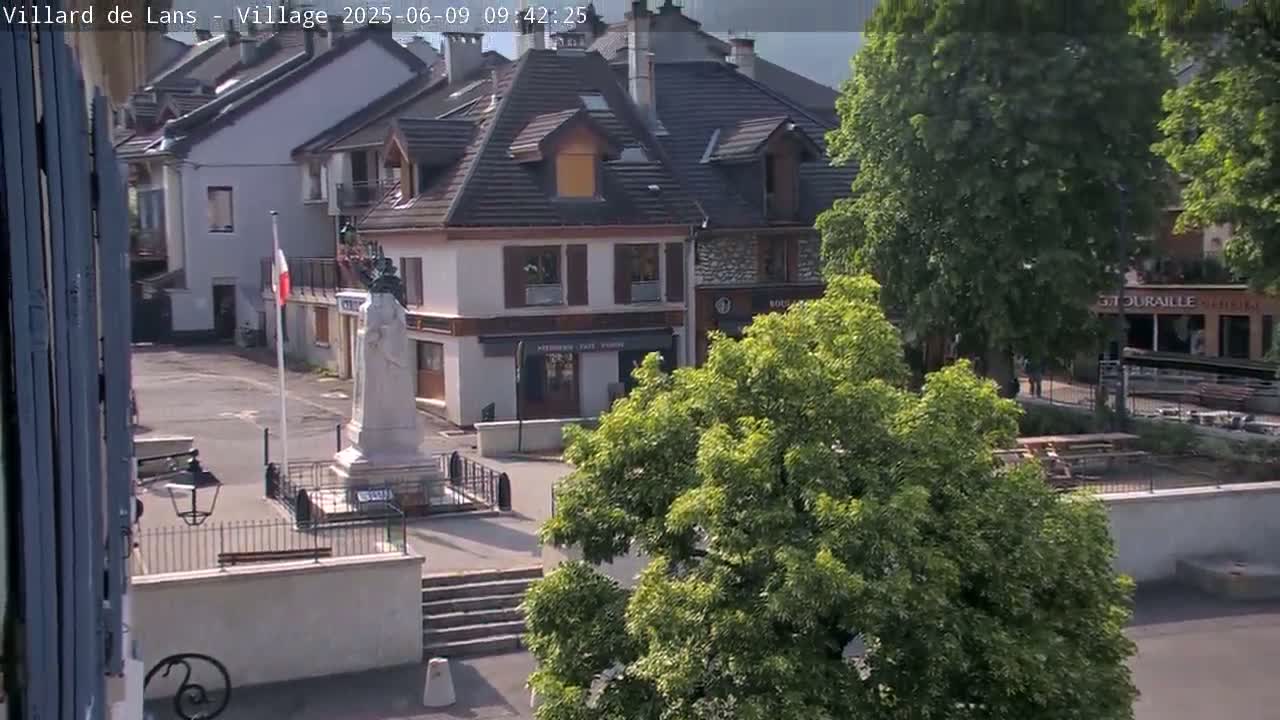 A mostly sunny view of a town square with a statue, buildings, and lush green trees.
