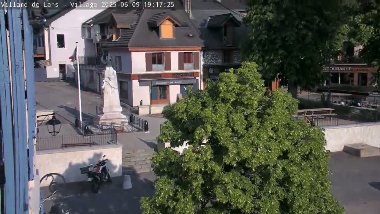A town square with a war memorial, several buildings, a motorcycle parked nearby, and a large tree in the foreground on a sunny day.