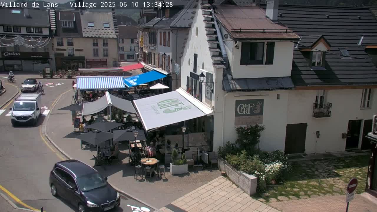A sunny village square with cars parked along the street, outdoor seating areas at cafés, and pedestrians walking nearby.