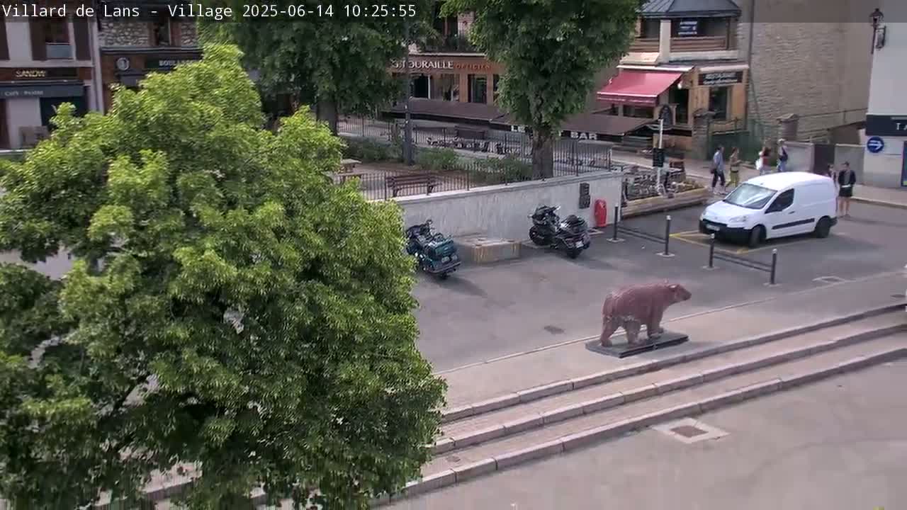 A sunny outdoor scene shows a village square with a large tree, several parked motorcycles, a white van, a statue of a bear, and several people standing near a building.