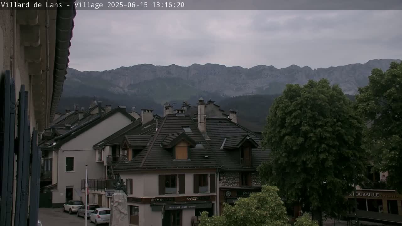 A village of buildings with dark-tiled roofs sits before a mountain range under an overcast sky.