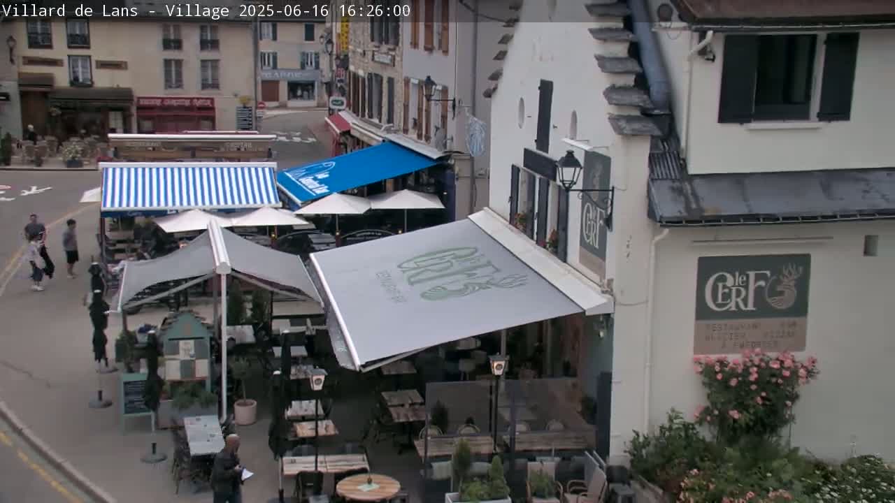 A street-level view shows an outdoor café with several striped and solid-colored awnings shading tables and chairs, situated in front of buildings on a sunny day.