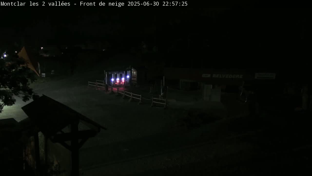 A nighttime, dark view of a mostly empty outdoor area with a small building lit with colored lights and a low fence.