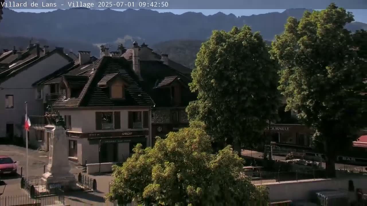 A village square with a monument, several buildings, and large trees under a sunny sky with mountains in the background.