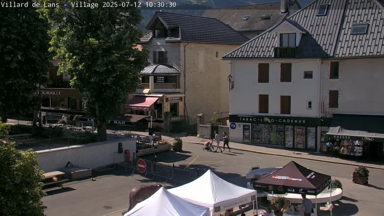 A sunny town square with several buildings, some outdoor seating areas, and people walking, with market stalls being set up.