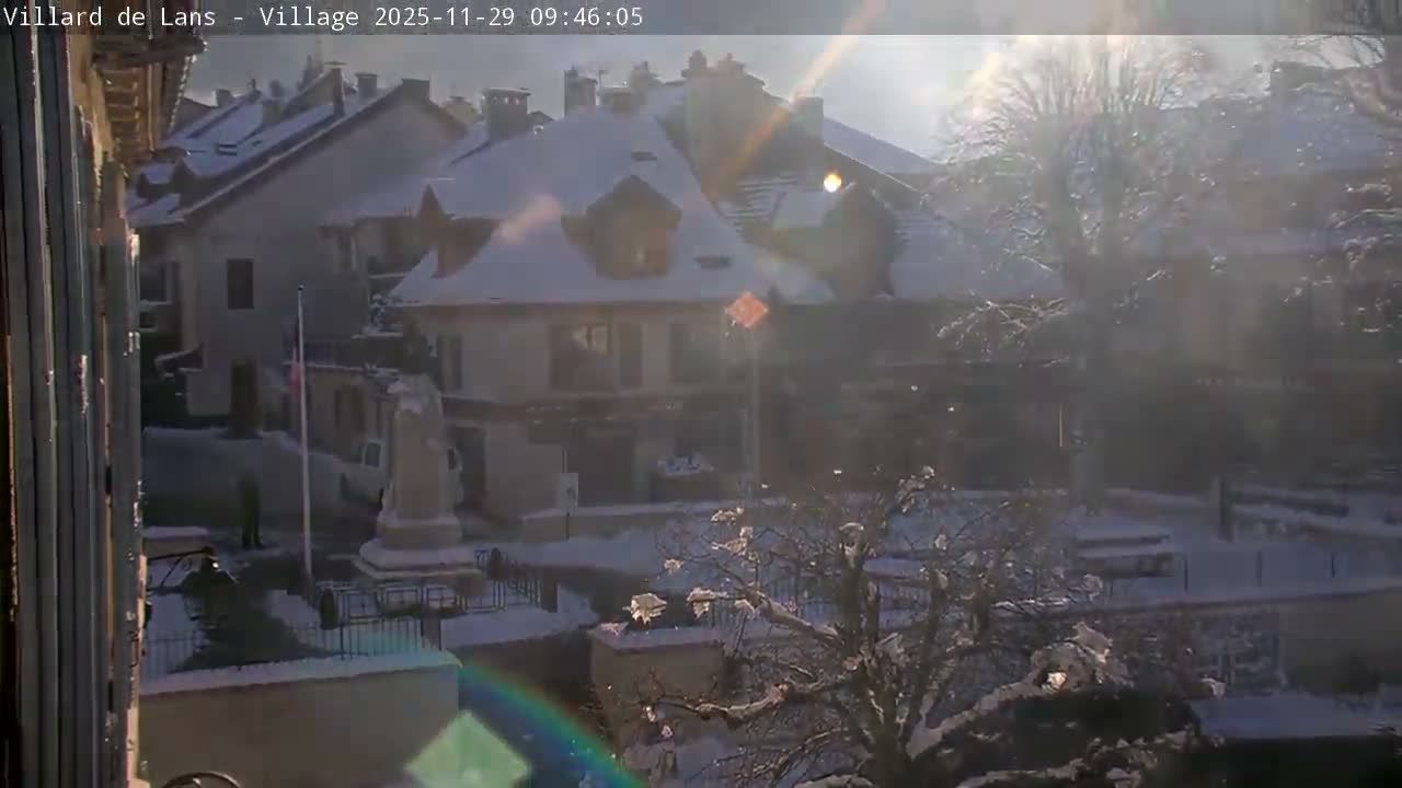 A bright, sunny day illuminates a snow-covered village, revealing rooftops draped in white, bare trees, and a central square featuring a statue and flagpole.