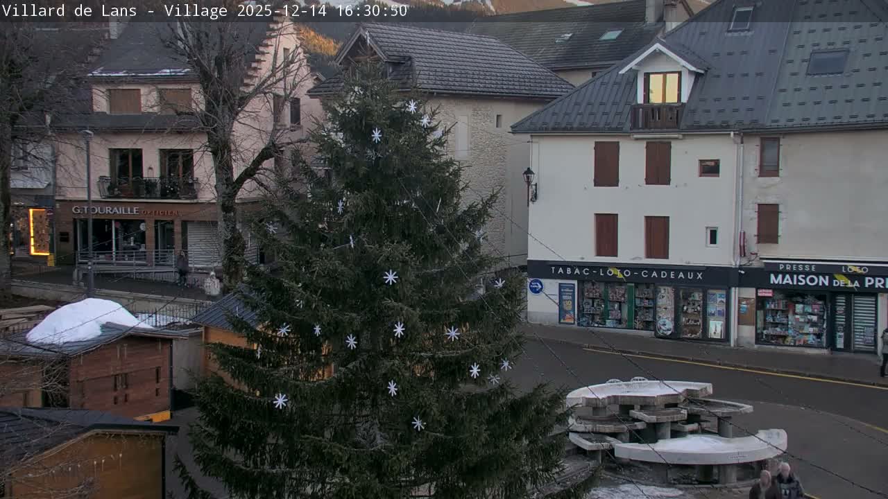 A snowy village square features a large, decorated Christmas tree, several snow-covered wooden market stalls, and a tiered stone picnic area, all under a steady fall of snow.