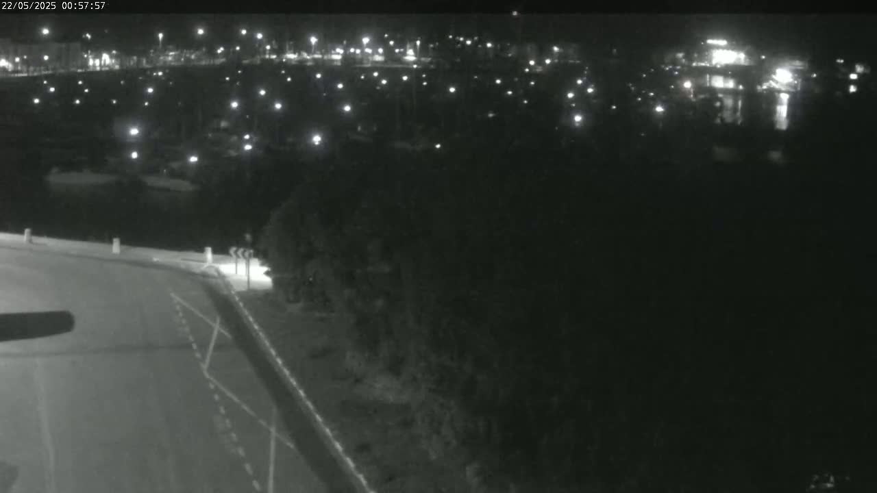 A nighttime, low-angle view shows a dark road curving to the right, leading toward a body of water dotted with small lights from boats, and a dark shoreline beyond.