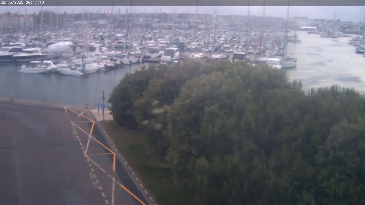 A harbor filled with numerous boats and yachts is visible from an elevated vantage point, with overcast skies and a dark-colored road in the foreground.
