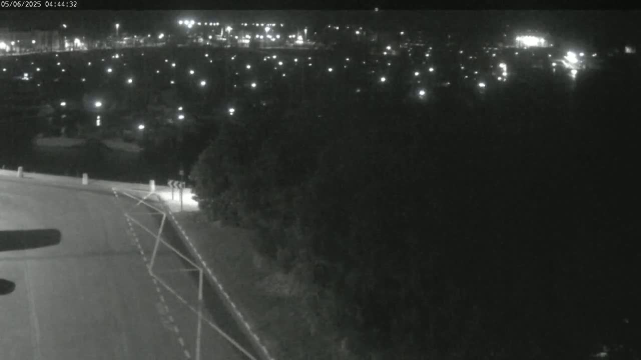 A nighttime view shows a dark road curving to the right, leading towards a distant, illuminated cityscape partially obscured by trees.