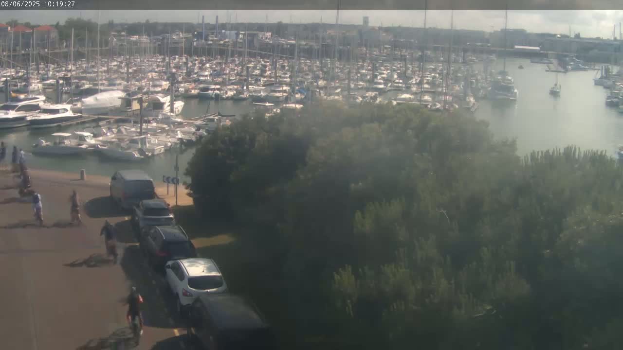 A marina filled with numerous boats is visible from above, with a hazy or slightly foggy atmosphere, and several people are biking along a road next to a line of parked cars.