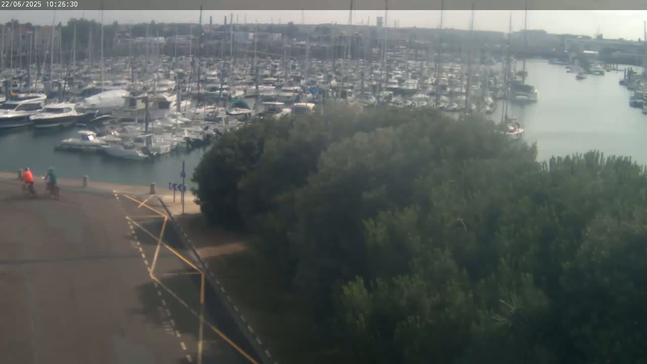 Two cyclists ride along a paved path overlooking a marina filled with numerous boats on a hazy day.