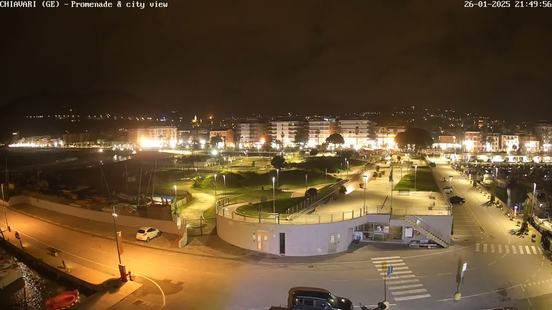 An illuminated nighttime view of a harbor promenade and city, featuring a landscaped park, parked cars, and boats in the water.