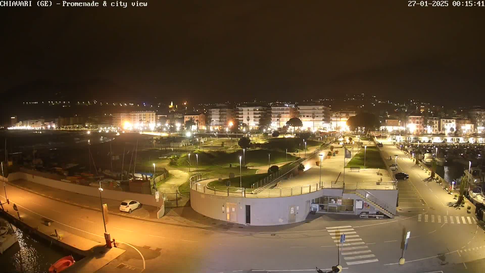 A nighttime aerial view shows a brightly lit promenade and harbor area next to a city, under clear skies.