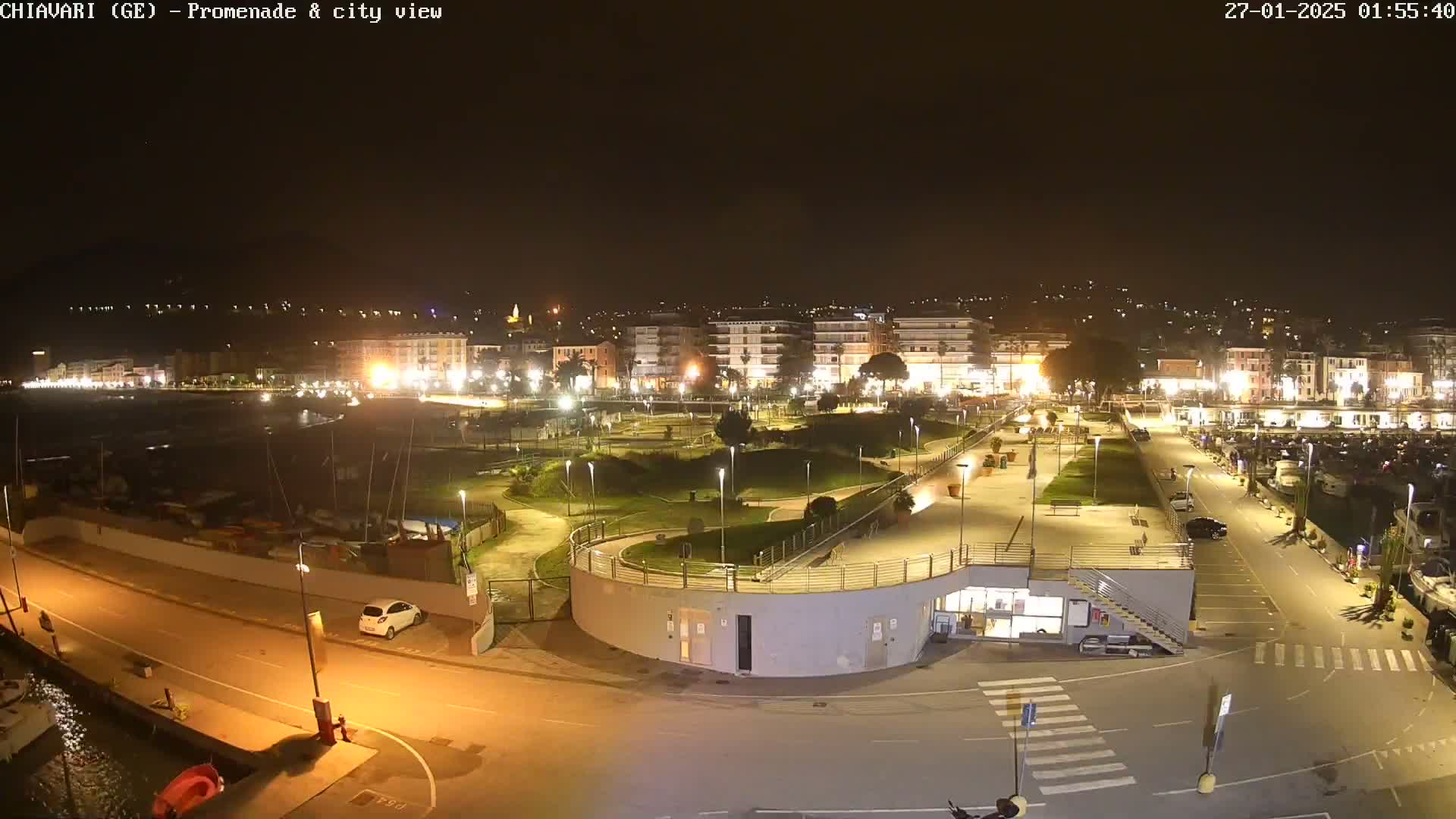 A nighttime view of a city's waterfront promenade and marina, showing a park-like area with walkways and buildings illuminated by streetlights.