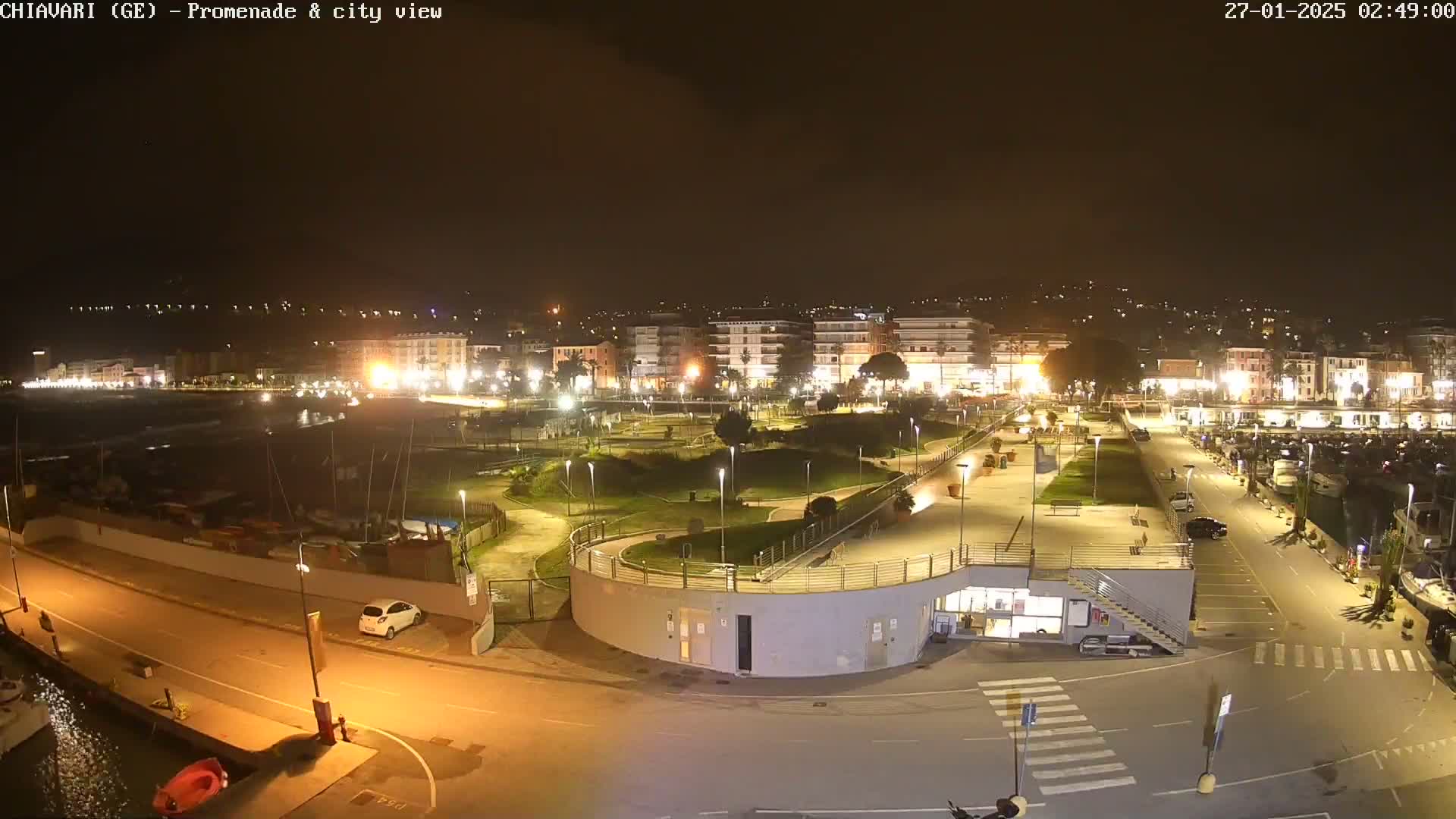 A nighttime, aerial view shows a brightly lit city promenade and harbor with a marina, park, and a few cars.