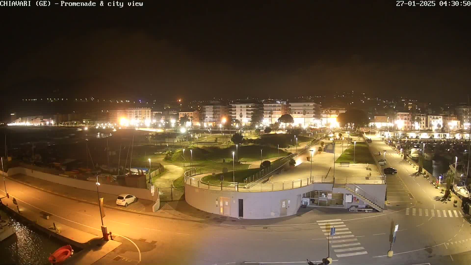 A nighttime view of a city's illuminated promenade and harbor, with boats moored and cars parked.